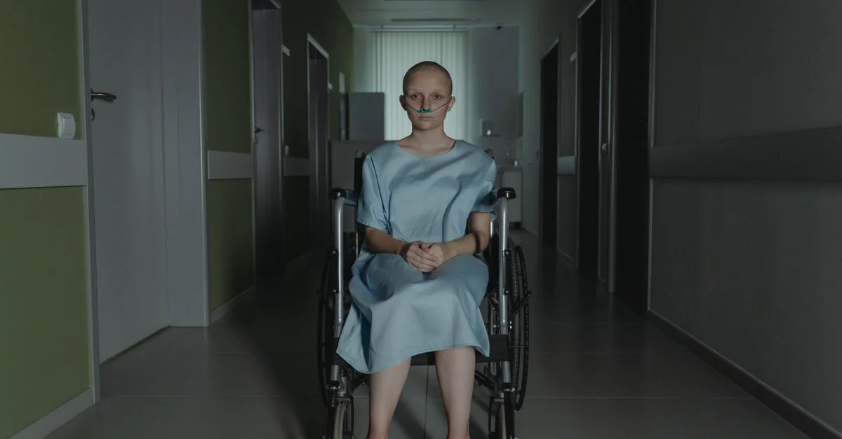 Une jeune femme en fauteuil roulant est assise dans un couloir d'hôpital, vêtue d'une robe de chambre, évoquant des thèmes de santé et de résilience.
