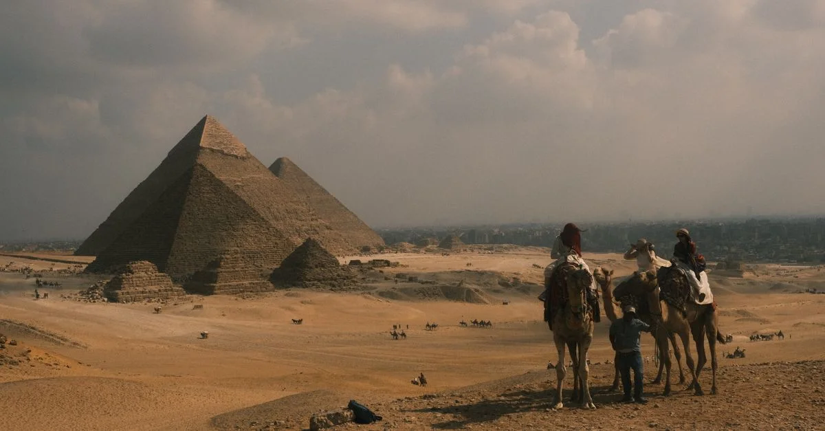 Des cavaliers à dos de chameau traversent le désert près des emblématiques pyramides de Gizeh sous un ciel nuageux.