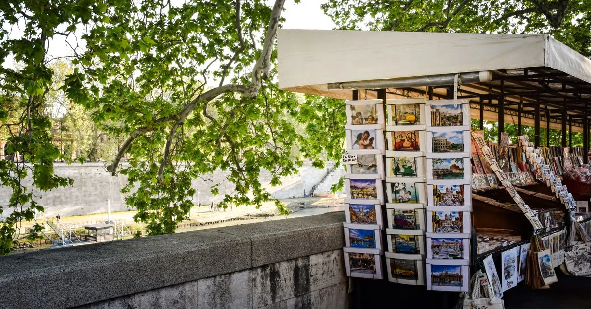 Stand de souvenirs à Rome, en Italie, présentant des cartes postales et des sacs sous des arbres luxuriants.