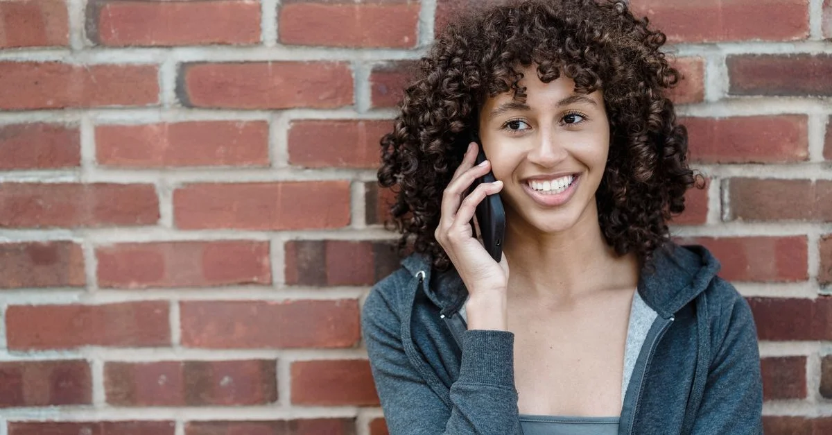 Femme joyeuse avec des cheveux bouclés utilisant un smartphone contre un mur en briques.