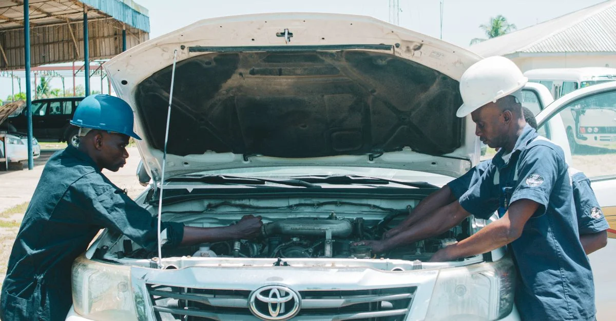 Deux mécaniciens auto au Nigeria travaillant sur un moteur de voiture en plein air, mettant en avant le travail d'équipe dans la réparation de véhicules.