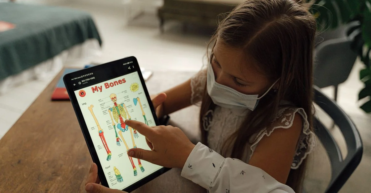 Une jeune fille portant un masque utilise une tablette pour étudier l'anatomie à la maison, mettant en avant l'apprentissage pendant la pandémie.